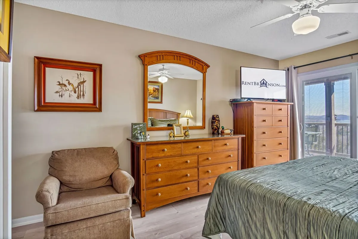 A cozy bedroom with a decorative bedspread, dresser with mirror, comfortable chair, and a TV on the dresser. The space feels welcoming, ideal for unwinding after a long day.