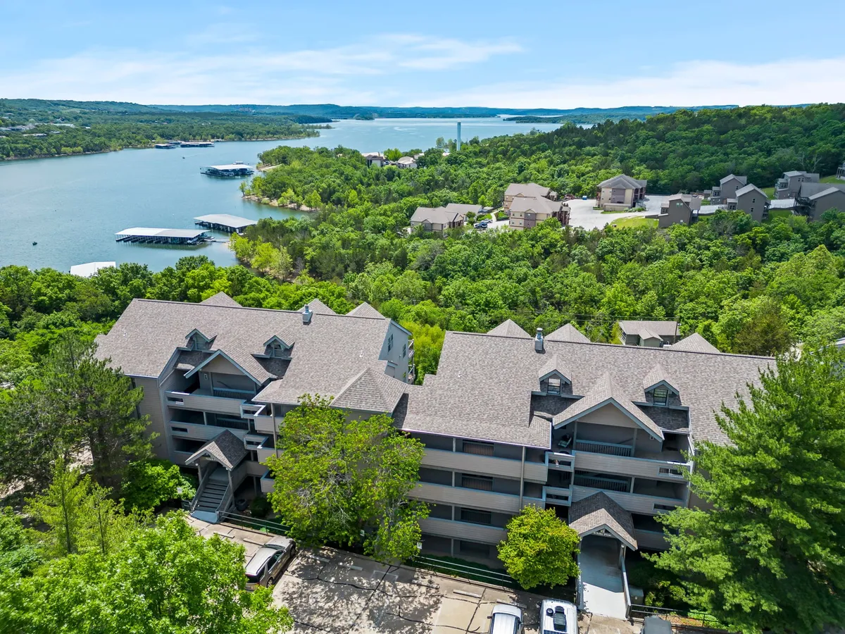 A wide exterior view showing a multi-story resort building nestled among lush greenery, with the lake visible in the foreground and distant hills creating a scenic backdrop. The setting radiates tranquility and resort living.
