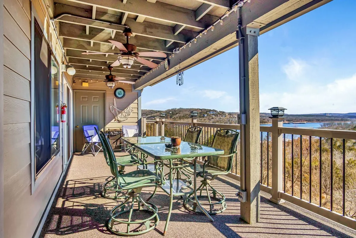 A private deck with a glass table, green chairs, scenic lake view, and ceiling fans. Designed for outdoor dining, relaxing, and soaking in the breeze, the deck offers a tranquil space just outside the main living area.