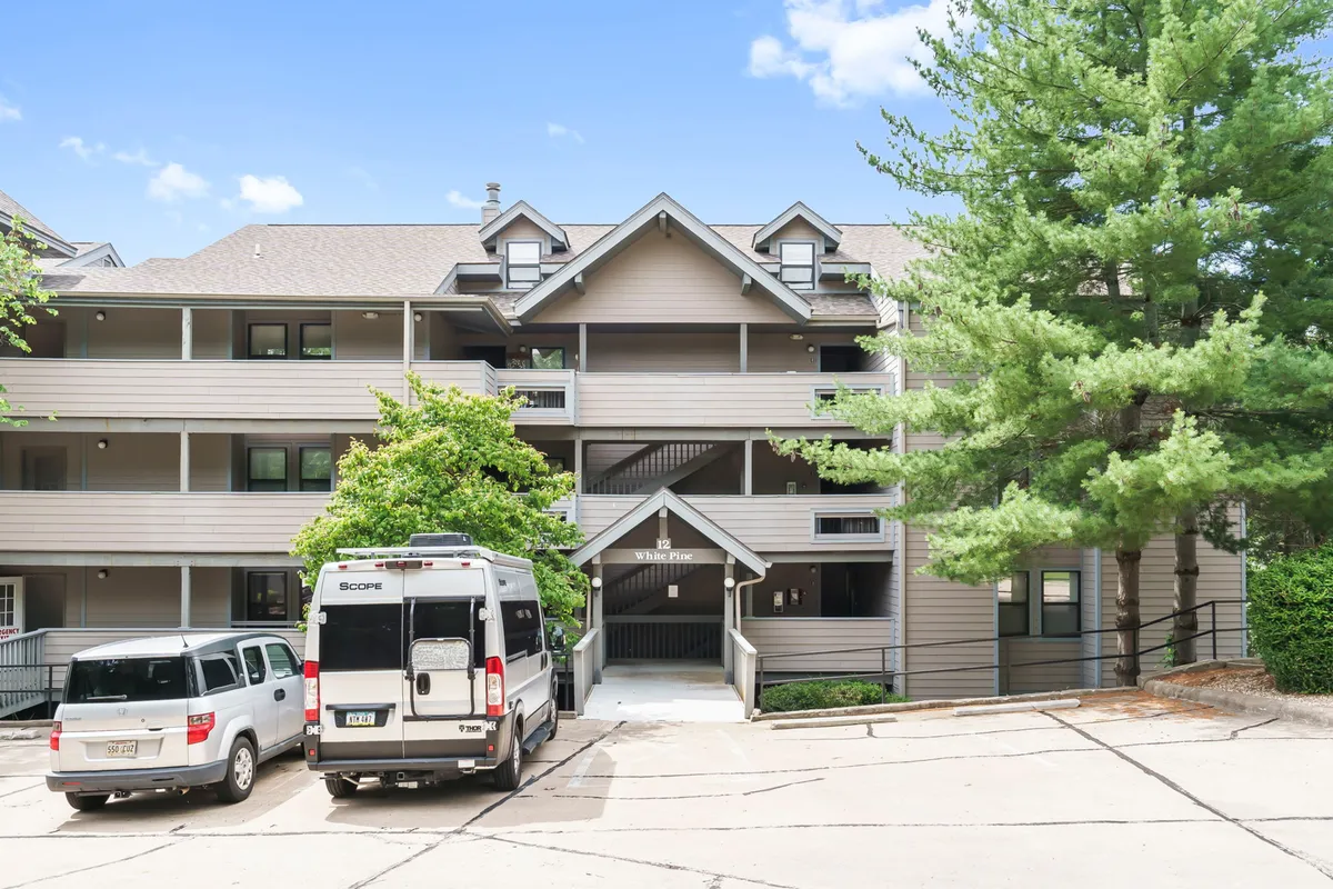 The building’s welcoming facade framed by leafy green trees, with balconies visible and vehicles parked nearby. The ambiance is calm and inviting, hinting at comfortable outdoor spaces and scenic views from within.