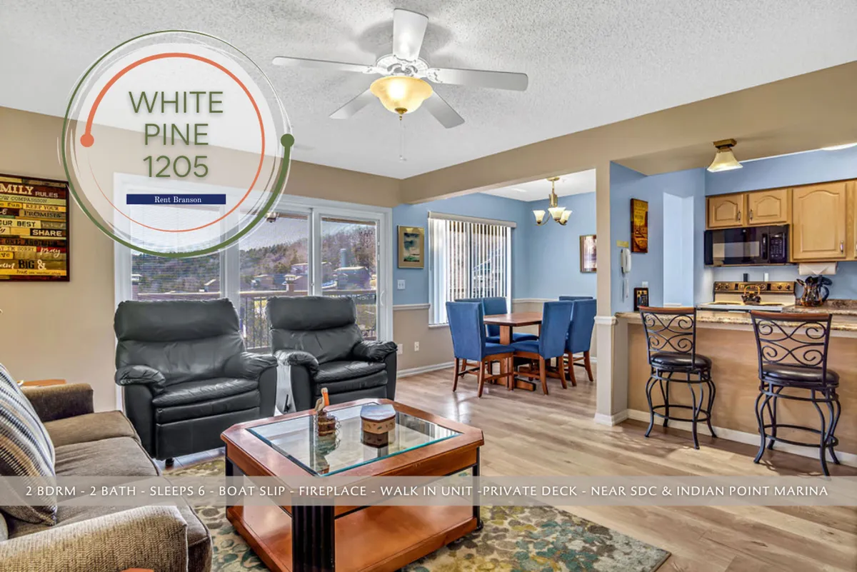 A spacious and sunlit open-concept living and dining area featuring comfortable leather chairs, a wooden dining table, a kitchen island with bar stools, and a decorative fireplace. Large windows allow natural light to pour in, and the overall atmosphere feels welcoming and homely.