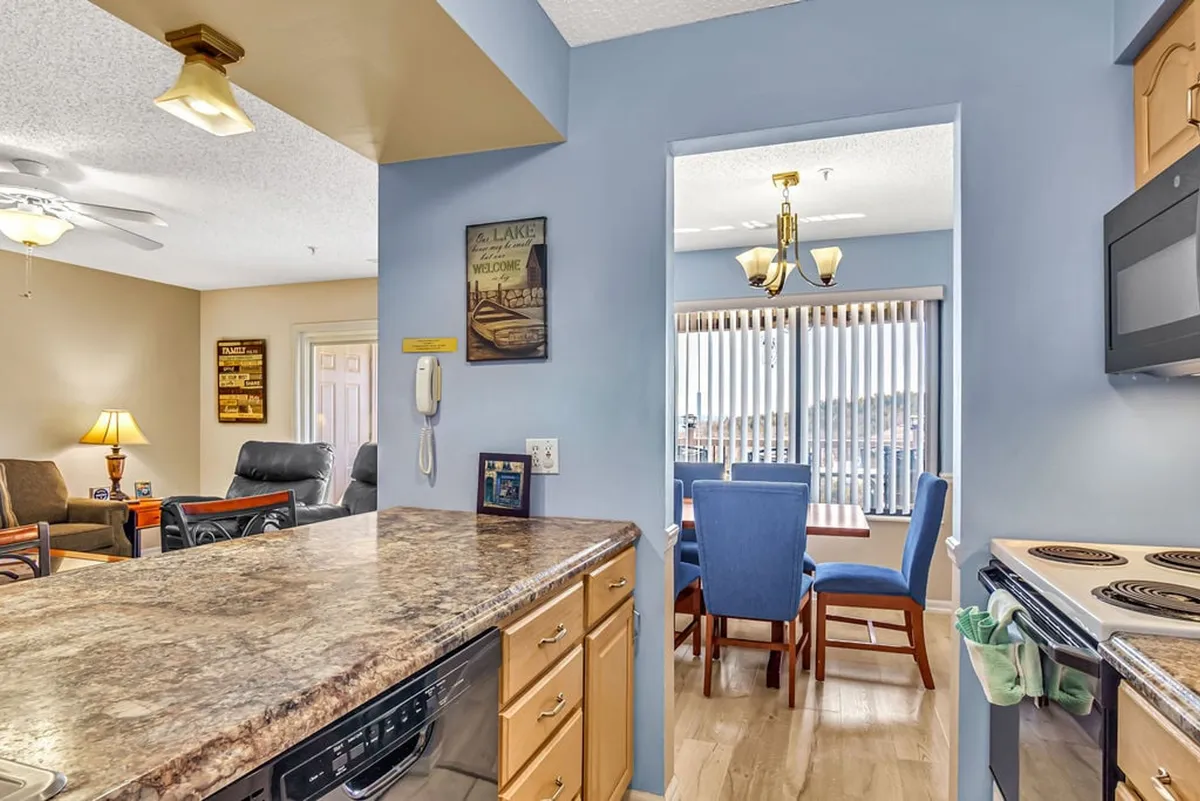 A stylish kitchen and dining area with a granite-topped kitchen island, blue dining chairs, and wall decor featuring a lake theme. The space is open and inviting, perfect for shared meals and family gatherings.